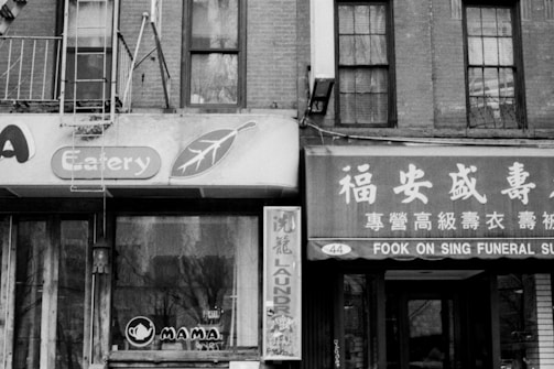 A black and white photograph of a city street view showcasing buildings with signage for an eatery and a funeral supply shop. The architecture is typical of older urban areas, with brick facades and fire escapes.