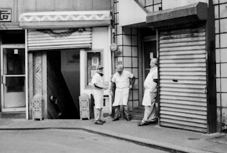 Three individuals in aprons stand and converse near the entrance of a building with closed metal shutters. The scene is urban, likely outside a shop, showcasing a casual interaction between workers during a break.