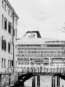 A large cruise ship named Viking is situated behind a traditional European-style building. A wooden bridge crossing a waterway is visible in the foreground, with a few people standing on it. The image is in black and white, highlighting the architectural contrast between the modern ship and the historic building.