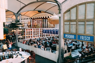 A bustling restaurant interior with many people seated at tables, engaged in conversation and dining. The decor is vintage with high ceilings and large windows, and there are shelves filled with various bottles behind the seating area. A large decorative fish hangs from the ceiling near a sign that reads 'Pescado Fresco.' The ambiance is lively and social.