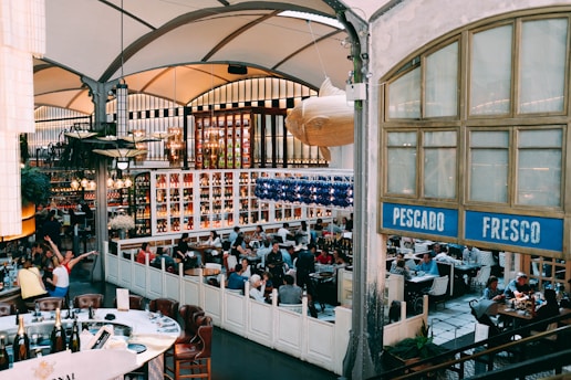 A bustling restaurant interior with many people seated at tables, engaged in conversation and dining. The decor is vintage with high ceilings and large windows, and there are shelves filled with various bottles behind the seating area. A large decorative fish hangs from the ceiling near a sign that reads 'Pescado Fresco.' The ambiance is lively and social.