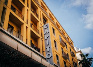 A multi-story yellow building with numerous balconies has a vertical sign reading 'CAPITOL' attached to its facade. The architectural style appears modern with clean, straight lines. A blue sky with scattered clouds forms the backdrop, and part of a palm tree is visible at the bottom right corner.