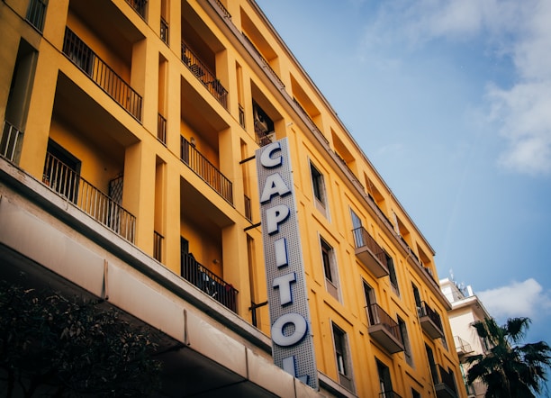 A multi-story yellow building with numerous balconies has a vertical sign reading 'CAPITOL' attached to its facade. The architectural style appears modern with clean, straight lines. A blue sky with scattered clouds forms the backdrop, and part of a palm tree is visible at the bottom right corner.