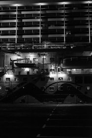 A large ship is docked, displaying multiple decks with balconies. The scene is dark, illuminated by a few spotlights highlighting parts of the ship's structure. The architecture suggests a cruise ship with evenly spaced windows and lifeboats on the deck.