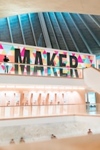 A modern indoor space featuring a large, colorful sign with the word 'MAKER' in bold letters against a backdrop of colorful geometric patterns. The architectural design includes a high ceiling with vertical beams and multiple floors visible. The area below the sign looks well-lit with displays and presentations visible in the background. Few people are present, some are standing and others are sitting.