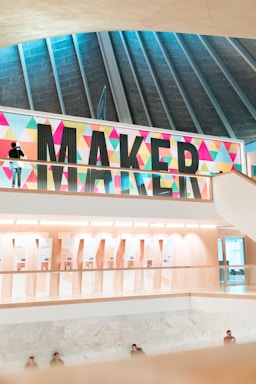 A modern indoor space featuring a large, colorful sign with the word 'MAKER' in bold letters against a backdrop of colorful geometric patterns. The architectural design includes a high ceiling with vertical beams and multiple floors visible. The area below the sign looks well-lit with displays and presentations visible in the background. Few people are present, some are standing and others are sitting.