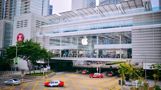 A modern, large glass-fronted building with a prominent Apple logo, housing an Apple Store. The structure is surrounded by high-rise buildings and features a busy street with several red taxis and cars. Trees and road signage appear in the foreground.