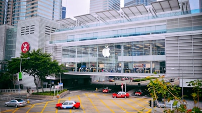 A modern, large glass-fronted building with a prominent Apple logo, housing an Apple Store. The structure is surrounded by high-rise buildings and features a busy street with several red taxis and cars. Trees and road signage appear in the foreground.