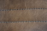 A close-up view of a brown leather surface with two parallel rows of dark stitching running horizontally across it. The texture of the leather is visible, with slight imperfections and a matte finish. The stitching appears uniform and evenly spaced.