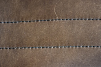 A close-up view of a brown leather surface with two parallel rows of dark stitching running horizontally across it. The texture of the leather is visible, with slight imperfections and a matte finish. The stitching appears uniform and evenly spaced.