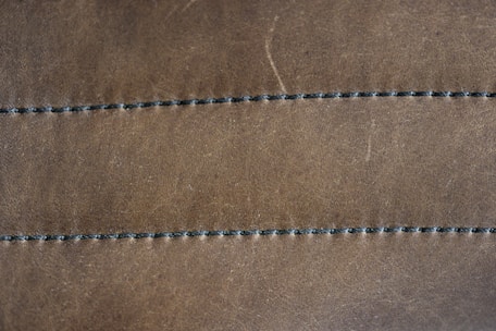 A close-up view of a brown leather surface with two parallel rows of dark stitching running horizontally across it. The texture of the leather is visible, with slight imperfections and a matte finish. The stitching appears uniform and evenly spaced.