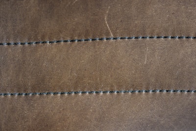 A close-up view of a brown leather surface with two parallel rows of dark stitching running horizontally across it. The texture of the leather is visible, with slight imperfections and a matte finish. The stitching appears uniform and evenly spaced.