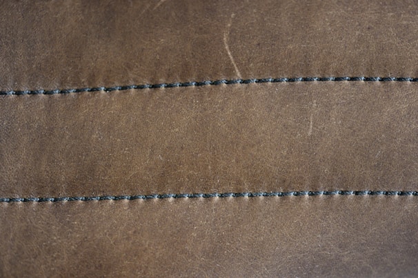 A close-up view of a brown leather surface with two parallel rows of dark stitching running horizontally across it. The texture of the leather is visible, with slight imperfections and a matte finish. The stitching appears uniform and evenly spaced.
