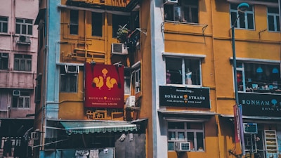 A vibrant yellow building with multiple windows and various signs, some with the name 'Bonham Strand'. Air conditioning units are visible, and there's a small green awning on the lower left. The structure shows signs of urban life, with plants and other personal items visible through the windows.