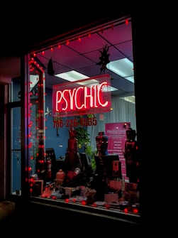 A storefront features a prominently displayed neon sign with the word 'PSYCHIC' in bright red. The window is adorned with red string lights and contains various decorative items, including statues and plants. A poster inside provides additional information on psychic services.
