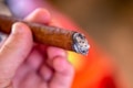 A hand is holding a partially smoked cigar with ash at the burning end. The cigar is brown with a visible texture on the wrapper. The background is blurred in warm tones, highlighting the focus on the hand and the cigar.
