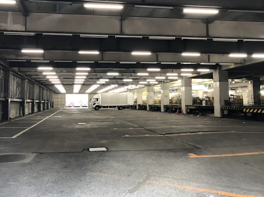An indoor loading dock area with a large, open space. Fluorescent lights are affixed to the ceiling, illuminating the area. A white delivery truck is parked on the right side near a section with loading bays. The area appears industrial, with exposed concrete floors and columns.