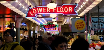 A bustling market scene with neon signs hanging from the ceiling indicating 'Lower Floor' and 'Market Grill Seafood'. Numerous people are walking through the area, browsing various stalls selling fresh produce and goods with vibrant displays of fruits, vegetables, and other items.