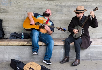 Two musicians are seated on a wooden bench against a concrete wall. The musician on the left plays an acoustic guitar and appears to be singing passionately, while the one on the right plays a banjo and is wearing a hat and sunglasses. A guitar case with a tip sign is on the ground in front of them.