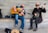 Two musicians are seated on a wooden bench against a concrete wall. The musician on the left plays an acoustic guitar and appears to be singing passionately, while the one on the right plays a banjo and is wearing a hat and sunglasses. A guitar case with a tip sign is on the ground in front of them.