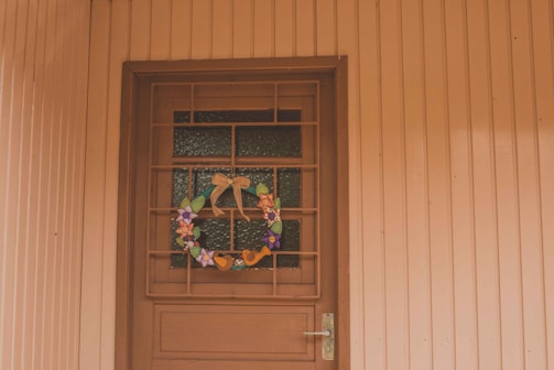 A wooden door set within a paneled wall is adorned with a decorative wreath. The wreath includes a variety of colorful flowers and ribbons, adding a touch of festivity. The light hue of the wood gives a warm and welcoming appearance.