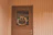 A wooden door set within a paneled wall is adorned with a decorative wreath. The wreath includes a variety of colorful flowers and ribbons, adding a touch of festivity. The light hue of the wood gives a warm and welcoming appearance.