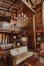 A rustic living room with log walls, a high ceiling, and a large chandelier. There is a beige sofa with visible wear, a wooden coffee table, and a variety of framed photographs and art pieces on the walls. A small balcony with a decorative railing is visible on the upper level.