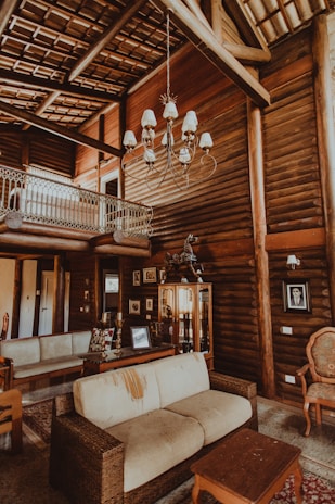 A rustic living room with log walls, a high ceiling, and a large chandelier. There is a beige sofa with visible wear, a wooden coffee table, and a variety of framed photographs and art pieces on the walls. A small balcony with a decorative railing is visible on the upper level.
