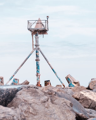 A weathered, graffiti-covered navigation structure stands on a rocky shore, supported by three angled metal legs. The structure features a triangular top and a small beacon, set against a clear blue sky.