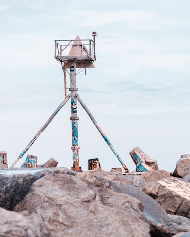 A weathered, graffiti-covered navigation structure stands on a rocky shore, supported by three angled metal legs. The structure features a triangular top and a small beacon, set against a clear blue sky.