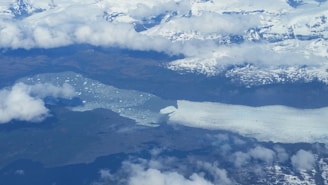 An aerial view of a large glacier extending into a lake or sea. The landscape is covered with snow and ice, with some dark areas of exposed earth. Clouds are scattered across the sky, giving the scene a cold and remote atmosphere.