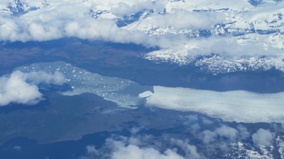 An aerial view of a large glacier extending into a lake or sea. The landscape is covered with snow and ice, with some dark areas of exposed earth. Clouds are scattered across the sky, giving the scene a cold and remote atmosphere.