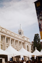 A historic building with classic architectural features such as columns and a prominent white steeple is in the background. In the foreground, several white tents are set up, possibly for an event. There are groups of people beneath the tents.