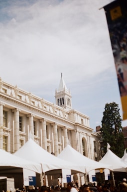 A historic building with classic architectural features such as columns and a prominent white steeple is in the background. In the foreground, several white tents are set up, possibly for an event. There are groups of people beneath the tents.