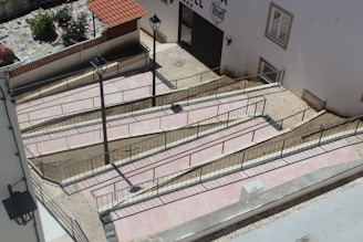 A series of interconnected ramps with railings are constructed against a building's exterior. The ramps are paved with light-colored tiles, bordered by metal railings for safety. Adjacent to the ramps, two street lamps are mounted, providing illumination. A small section of a garden with flowers and a tiled area is visible in the background, enclosed by a low wall with a red tiled roof.
