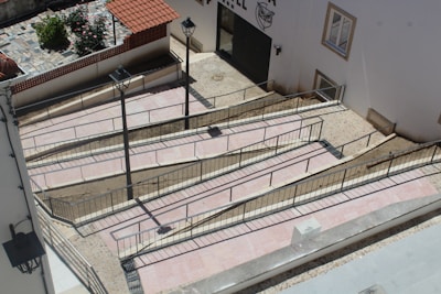 A series of interconnected ramps with railings are constructed against a building's exterior. The ramps are paved with light-colored tiles, bordered by metal railings for safety. Adjacent to the ramps, two street lamps are mounted, providing illumination. A small section of a garden with flowers and a tiled area is visible in the background, enclosed by a low wall with a red tiled roof.