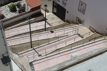 A series of interconnected ramps with railings are constructed against a building's exterior. The ramps are paved with light-colored tiles, bordered by metal railings for safety. Adjacent to the ramps, two street lamps are mounted, providing illumination. A small section of a garden with flowers and a tiled area is visible in the background, enclosed by a low wall with a red tiled roof.