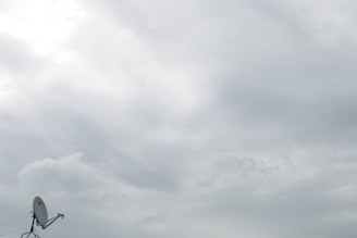 A cloudy sky dominates the view, with thick layers of gray clouds covering most of the area. In the lower left corner, a satellite dish is visible, mounted on a rooftop, pointing upwards towards the clouds.