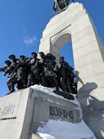 A large war memorial made of stone and metal features soldiers in uniform marching forward, set beneath a grand arch. The years 1939-1945 and 1914-1918 are inscribed, indicating world war dates. Snow is visible on the structure, with clear blue skies in the background.