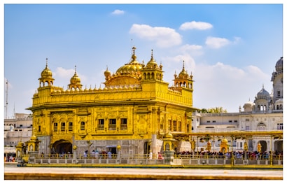 A magnificent golden temple with intricate architectural details surrounded by a clear blue sky. The structure features ornate decorations and is a prominent focal point, attracting visitors who are gathered around the perimeter.