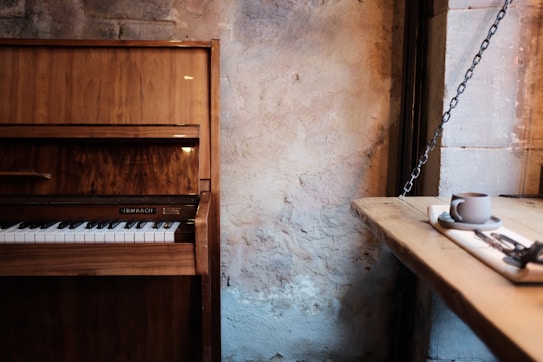 An upright wooden piano stands against a textured, rustic wall. A wooden table extends from the right side of the image, holding a coffee cup on a saucer with a napkin and cutlery, suggesting a cozy, vintage caf&eacute; setting.