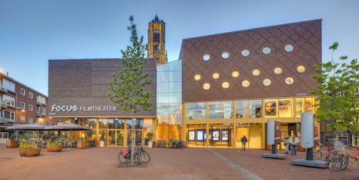 A modern building with a combination of brick and glass architecture, featuring numerous circular windows on the facade. The structure is identified as Focus Filmtheater with its sign visible on the front. Several bicycles are parked nearby, and there are a few people walking in the open plaza area in front. The sky is clear, suggesting daytime, and some green trees provide a touch of nature amidst the urban setting.