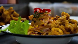 A plate of delicious-looking fried food garnished with fresh greens and sliced red peppers. The dish appears to be a mix of fried chicken or fish and thinly sliced vegetables. The background shows other plates of food out of focus.