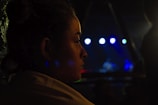 A dimly lit profile of a person looking towards a stage where a musical performance is taking place. Bright spotlights in shades of blue and white illuminate the background, creating a contrast with the dark surroundings. The person's face is partially illuminated, capturing their focus and contemplation.