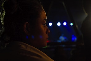 A dimly lit profile of a person looking towards a stage where a musical performance is taking place. Bright spotlights in shades of blue and white illuminate the background, creating a contrast with the dark surroundings. The person's face is partially illuminated, capturing their focus and contemplation.