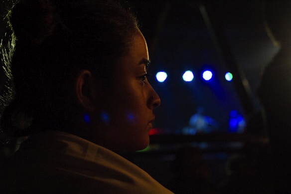 A dimly lit profile of a person looking towards a stage where a musical performance is taking place. Bright spotlights in shades of blue and white illuminate the background, creating a contrast with the dark surroundings. The person's face is partially illuminated, capturing their focus and contemplation.