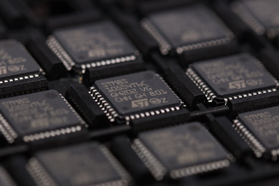 A close-up view of several integrated circuits or microchips arranged in rows. The surface details of the chips are visible, including markings such as part numbers and manufacturer logos. The chips are placed in a dark-colored tray designed for holding them securely.