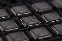 A close-up view of several integrated circuits or microchips arranged in rows. The surface details of the chips are visible, including markings such as part numbers and manufacturer logos. The chips are placed in a dark-colored tray designed for holding them securely.