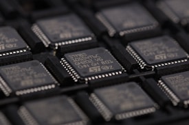 A close-up view of several integrated circuits or microchips arranged in rows. The surface details of the chips are visible, including markings such as part numbers and manufacturer logos. The chips are placed in a dark-colored tray designed for holding them securely.