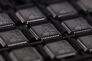 A close-up view of several integrated circuits or microchips arranged in rows. The surface details of the chips are visible, including markings such as part numbers and manufacturer logos. The chips are placed in a dark-colored tray designed for holding them securely.